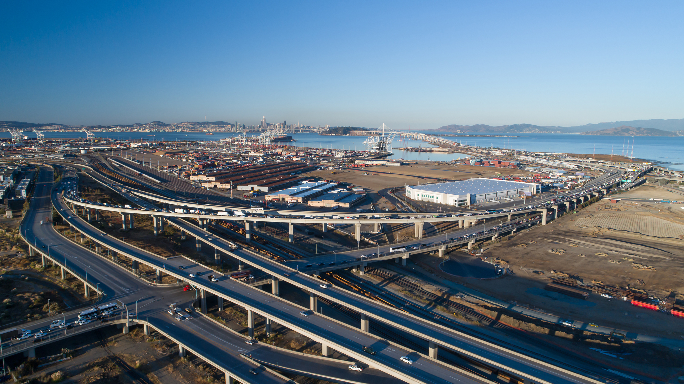 Aerial view of Prologis Oakland Global Logistics Center 2 in Oakland, California near busy highway with traffic. Showing bridge, port, the bay and city skyline in the background
