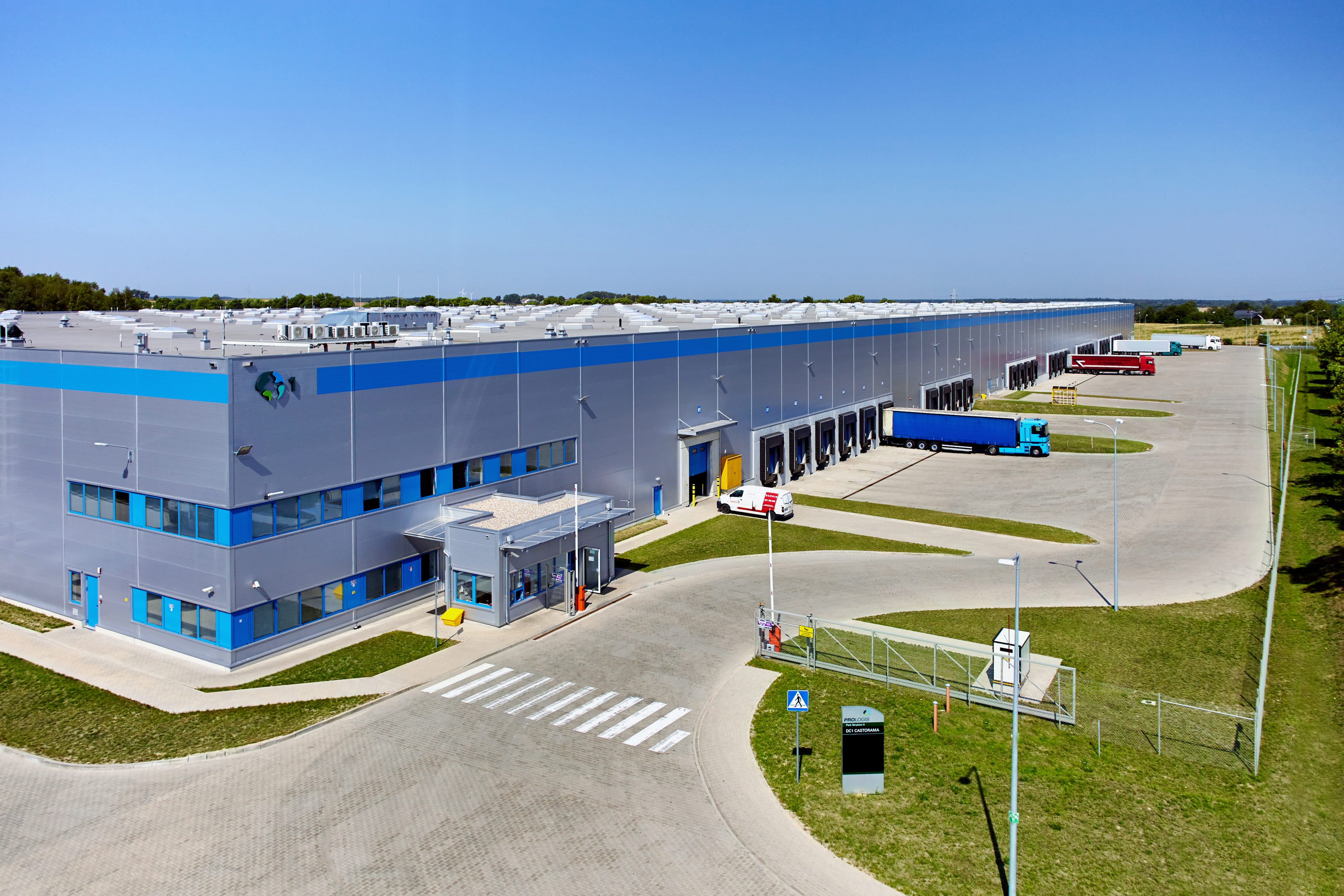 Angled view of warehouse loading dock at Prologis Park Strykow in Strykow, Poland. Blue stripe on building with Prologis globe and number 1, security gate and booth, trucks parked in lot and green lawn