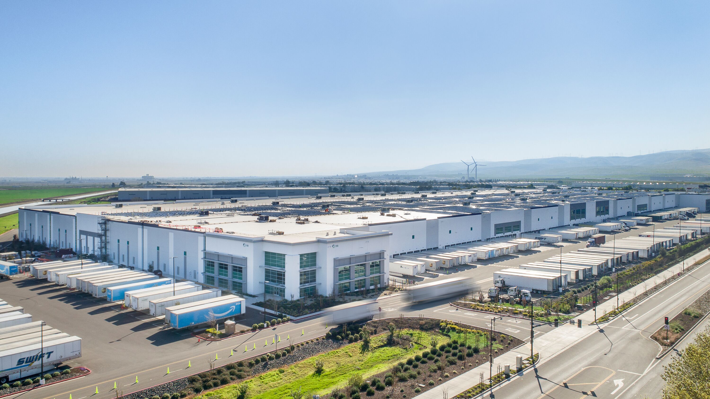 Exterior shot of Prologis International Park of Commerce in Tracy, California with semi trailers parked in loading docks, semi trucks driving in and out and windmills in the distance