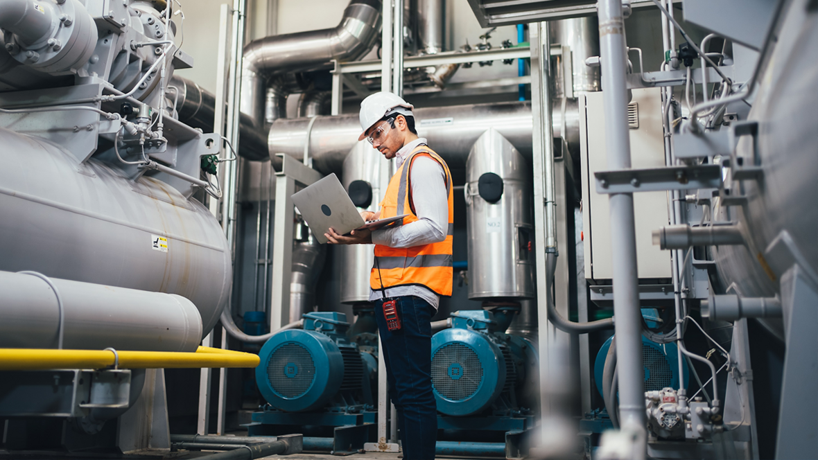 A photo of a man by machinery using a laptop