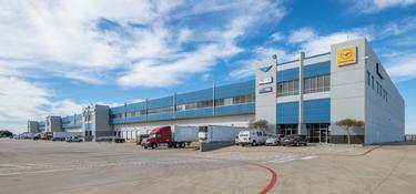 A photo of the truck court at Prologis DFW Air Cargo Center. The building is blue and grey with 4 air cargo logos shown on the side. trucks and cars are parked throughout the truck court