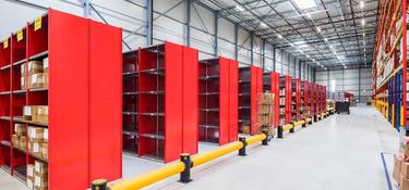A photo of the inside of Prologis Nieuwegein DC2. In the foreground there is short red racking holding cardboard boxes and in the background there is taller red and yellow racking.