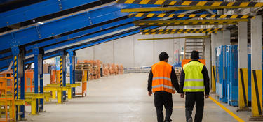 Two people walking inside of  a warehouse in orange and yellow construction vests, underneath blue and yellow conveyor racking