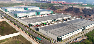 Angled aerial shot of Prologis Chongqing Liangjiang Logistics Center in China with industrial park in the distance, surrounding green space and trucks parked in loading area