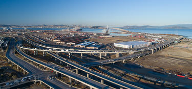 Aerial view of Prologis Oakland Global Logistics Center 2 in Oakland, California near busy highway with traffic. Showing bridge, port, the bay and city skyline in the background