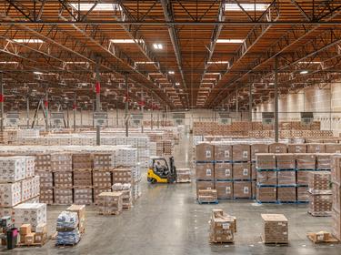 A photo of the interior of a warehouse with product stacked on pallets on the floor, and a yellow forklift driving in the aisles.