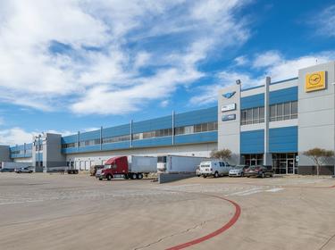 A photo of the truck court at Prologis DFW Air Cargo Center. The building is blue and grey with 4 air cargo logos shown on the side. trucks and cars are parked throughout the truck court