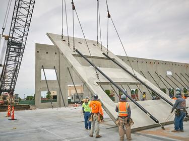 A photo of warehouse walls being stood up. contruction workers stand around the wall segment guiding it upwards in hard hards and green and orange work vests.
