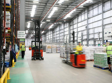 A photo of the interior of Prologis Coventry DC11. to the right there is product stacked on pallets on the floor, with red forklifts driving through a main aisle and to the left is tall racking. The warehouse is bustling with workers.