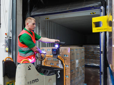A photo of a male worker in a green shirt and red vest on a worklift carrying pallets of product into a semi trailer using a scanner with a bright blue light.