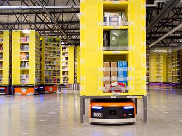 A photo small orange and black robots carrying stacks of products inside of a warehouse