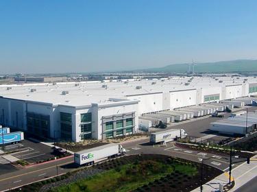 An exterior photo of Prologis Internation Park of Commerce. in the foreground are semi trailers parked in the truck court and in front of dock doors, and in the background are green hills and windmills.