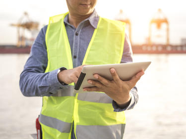 A photo of a person in a grey shirt and yellow construction vest holding an ipad with cranes in the background