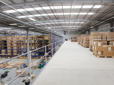 An interior photo of a warehouse, from the view of the mezzanine level. Below there are blue and yellow racks with product on the shelves