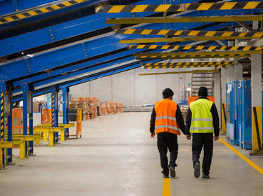 Two people walking inside of  a warehouse in orange and yellow construction vests, underneath blue and yellow conveyor racking