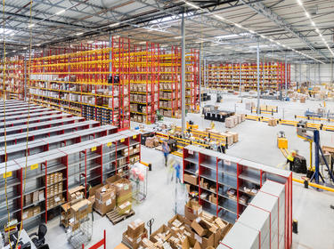 A busy interior of a warehouse, showing yellow and red racking, boxes stacked on the floor and workers moving around