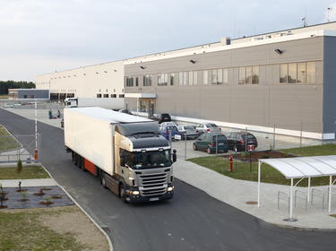 An exterior photo of a grey and white warehouse, with a truck driving out of the parking lot.