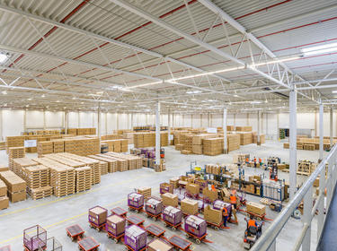 Interior warehouse angled view of Prologis Munich East Distribution Center 1 in Germany with stacked boxes and red and purple carts loaded. Multiple warehouse workers with orange vests
