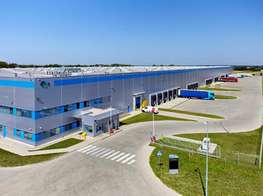 Angled view of warehouse loading dock at Prologis Park Strykow in Strykow, Poland. Blue stripe on building with Prologis globe and number 1, security gate and booth, trucks parked in lot and green lawn