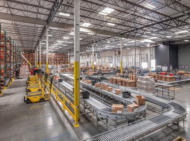Angled interior warehouse shot of Prologis Park Ontario in Ontario, California with parked forklifts lined up, conveyers running boxes, racking and bright LED lit interior.