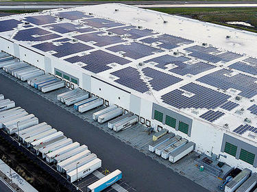 An aerial photo of Prologis Internaltional Park of Commerce. The roof of the building is covered in solar panels, and semi-truck trailers are parked in many of the dock doors in the foreground.  A grassy area is in the background.