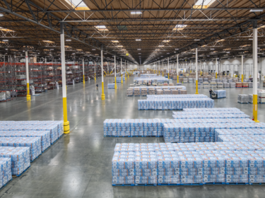 Interior warehouse wide angle shot of Prologis Kaiser Distribution Center 5 in Fontana, California with stacked crates of wrapped water bottles, a janitor, and woman driving forklift