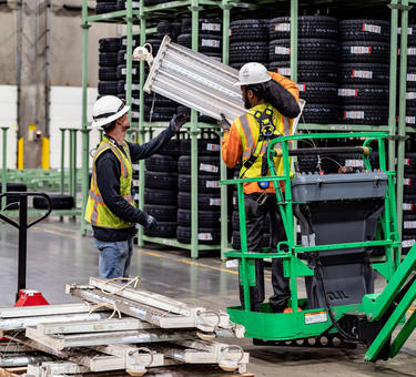 A photo of two men in a tire warehouse. On man holds an LED light fixture while standing on a green forkift, while the other guides it upwards