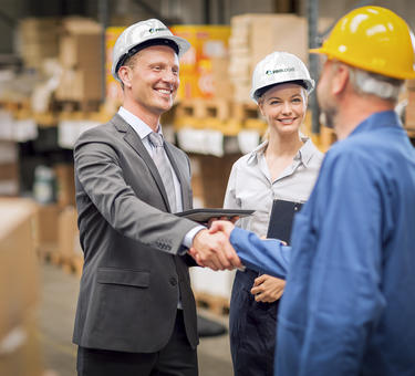 A smiling male Prologis team member in a hard hat shakes hands with a warehouse worker while female employee in hard hat stands to the right